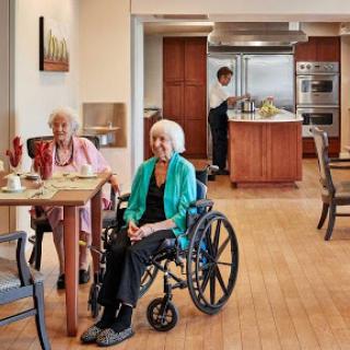 Two elderly women enjoy their time in a dining area, while a caregiver prepares food in the kitchen.