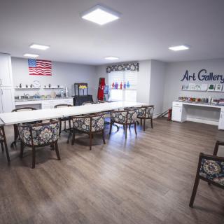A spacious art gallery interior with tables and chairs arranged for classes or workshops, featuring an American flag and art supplies on display.