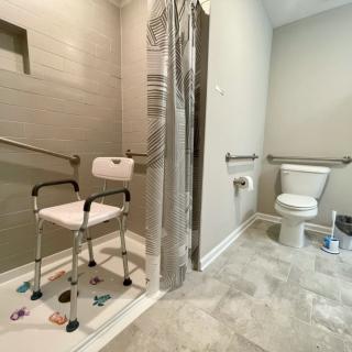 An accessible bathroom featuring a shower with a chair and grab bars, alongside a toilet equipped with safety handles.