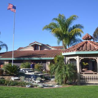 A scenic view of a gazebo and landscaped garden with a building and an American flag in the background under a clear blue sky.