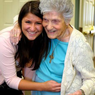 A young woman embraces an elderly woman, both smiling happily indoors.