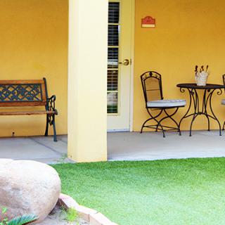 A cozy patio with a bench and a table set against a yellow wall, overlooking a patch of grass.