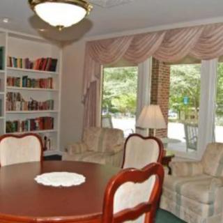 A cozy living room with a round table, surrounded by upholstered chairs, bookshelves filled with books, and large windows adorned with curtains.