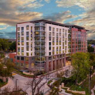 A modern multi-story building surrounded by trees and streets under a cloudy sky.