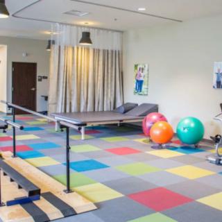 A well-equipped physical therapy room with colorful floor tiles and various exercise equipment, including balance bars and exercise balls.