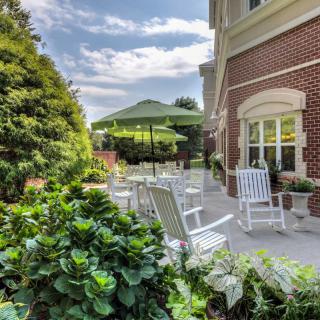 A serene garden patio with white rocking chairs and an umbrella, set against a brick building.