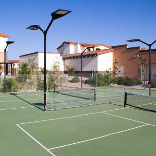 An outdoor tennis court surrounded by buildings and equipped with overhead lights, set under a clear blue sky.