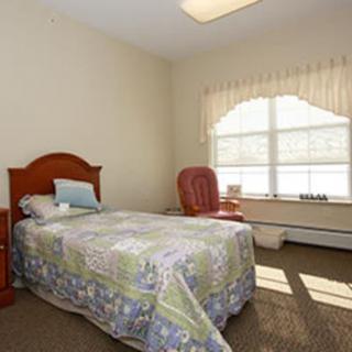 A small, cozy bedroom featuring a single bed with a colorful quilt, next to a large window with curtains. A comfortable chair is placed beside the window.