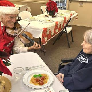 An elderly man in a Santa hat plays the violin for a woman during a festive holiday meal.