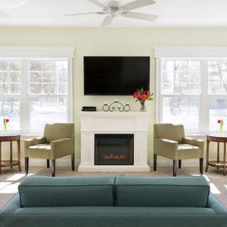 A modern living room features a fireplace with a mounted television above it, flanked by two armchairs and illuminated by natural light streaming through large windows.