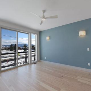 A bright and empty room with wooden flooring, blue accent wall, ceiling fan, and large windows opening to a balcony.