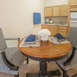 A cozy dining area with a wooden round table set for two, featuring decorative napkins and a flower arrangement in front of a kitchen space.