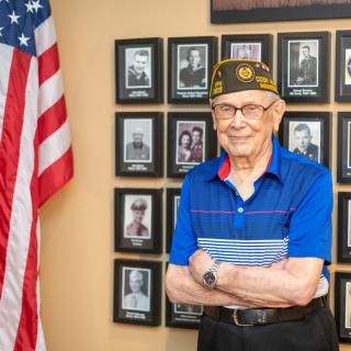 A veteran stands proudly in front of a memorial wall adorned with photos and an American flag.