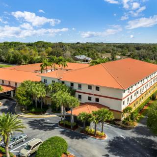 Aerial view of a large building with a red roof surrounded by trees and a parking lot.