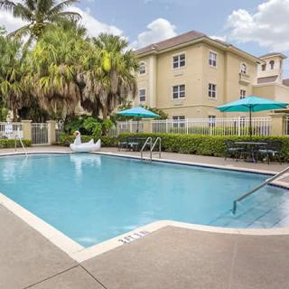 A tranquil outdoor swimming pool surrounded by palm trees and a building, featuring sun umbrellas for relaxation on a sunny day.