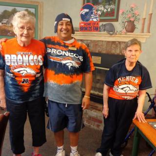 Three people wearing Broncos shirts pose for a group photo in a living room.