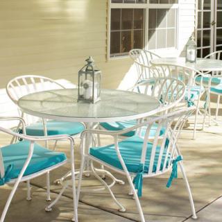 A sunlit patio with white metal furniture, featuring a round table and chairs accented with teal cushions, and a decorative lantern on the table.
