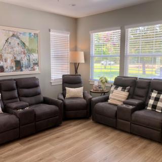 A cozy living room featuring recliner chairs and a sofa, with decorative pillows and a floor lamp by the windows. Artwork adorns the wall, creating a comfortable and inviting space.