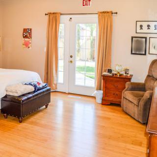 A cozy bedroom with wooden flooring, featuring a bed, armchair, and a side table next to large glass doors with curtains.