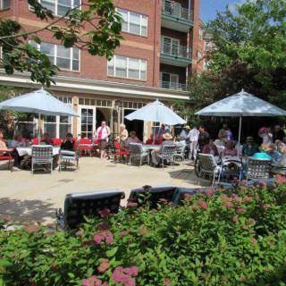A group of people enjoying a sunny day on a patio with umbrellas in front of a brick building.