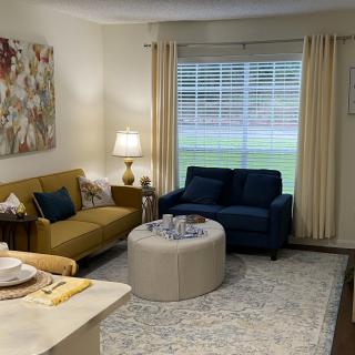 A cozy living room with a mix of modern and classic furniture, featuring a yellow and a blue sofa, an ottoman-style coffee table, and soft lighting from a table lamp.