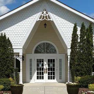 A white building entrance with double doors, surrounded by trees and plants.