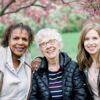 Three women of different generations smiling together in a garden with blooming trees.
