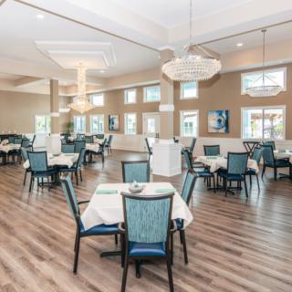 A modern dining room with neatly arranged tables and chairs, featuring elegant chandeliers and large windows.