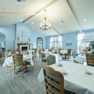 A spacious dining room with elegantly arranged tables and chairs, featuring a chandelier and large windows.