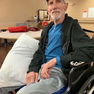 An elderly man sitting in a wheelchair, smiling and looking comfortable in a casual setting.