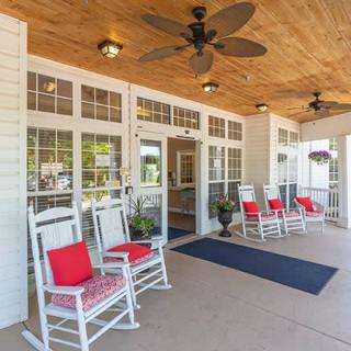 A cozy porch with white rocking chairs and red cushions, featuring ceiling fans and large windows at the entrance.