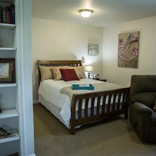 A cozy bedroom with a wooden bed, decorated pillows, a reclining chair, and a small bookshelf.