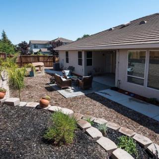 A residential backyard featuring a landscaped garden, patio, and outdoor seating area on a sunny day.