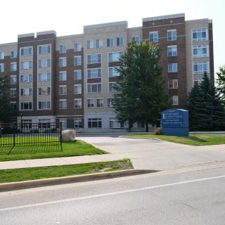 A multi-story residential apartment building with a sign and surrounding trees along a road.