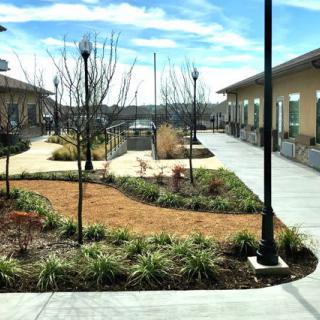 A landscaped courtyard featuring pathways and buildings, with lamp posts lining the area under a clear blue sky.