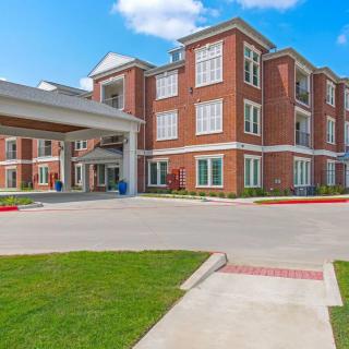 A modern brick apartment complex with a covered entrance and well-maintained lawn under a partly cloudy sky.