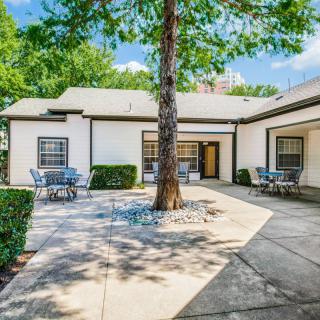 A tranquil courtyard featuring a large tree, surrounded by a building with tables and chairs under a clear sky.
