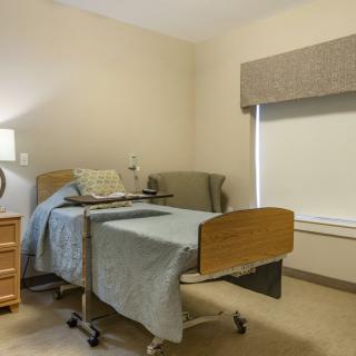 A clean and simple hospital room with a single bed, bedside table, and chair.