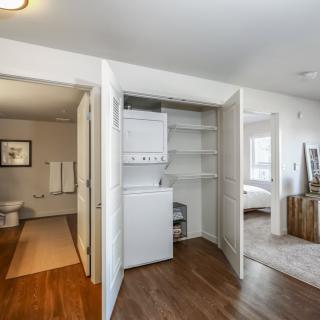 An apartment interior featuring a laundry area with a stacked washer and dryer, adjacent to a bathroom and bedroom.