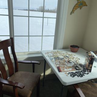 A cozy indoor setting with a puzzle spread out on a table near a window showing a snowy landscape outside.
