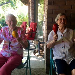 Two elderly women sitting together outdoors, smiling and holding maracas.