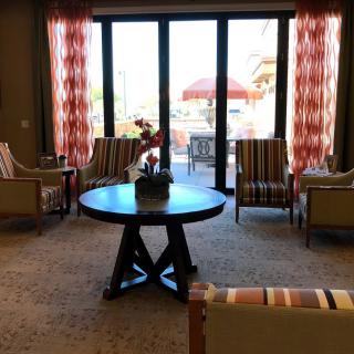 A cozy living room with a round table surrounded by chairs, featuring large windows and patterned curtains.