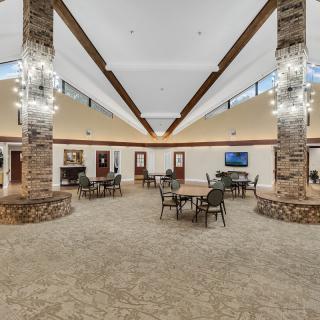 A spacious lounge area with stone pillars, chandeliers, and tables and chairs arranged on a carpeted floor.