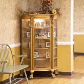A vintage curio cabinet filled with photographs, placed against a wallpapered wall next to a chair.