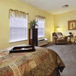 A cozy bedroom with warm lighting and classic decor, featuring a bed, armchair, lamp, and windows.