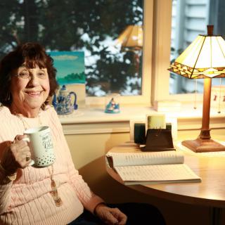A woman sitting at a desk holding a coffee mug, surrounded by a cozy atmosphere with a lamp and books.
