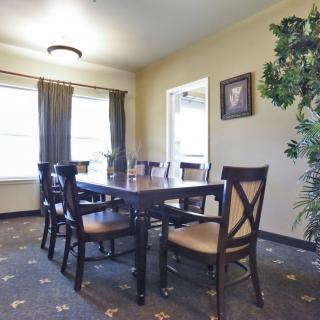 A well-lit dining room featuring a table with six chairs, large windows with curtains, and a patterned carpet.