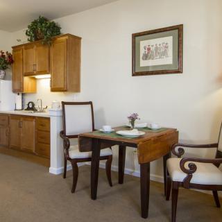 A cozy dining area with a small table and two chairs next to a kitchenette, adorned with wall art and subtle decor.