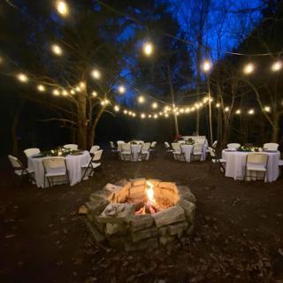 An enchanting outdoor evening event with beautifully arranged tables and a cozy firepit, illuminated by sparkling string lights.