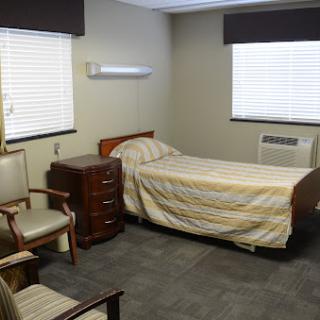 A simple room featuring a bed, chairs, and windows with blinds.
